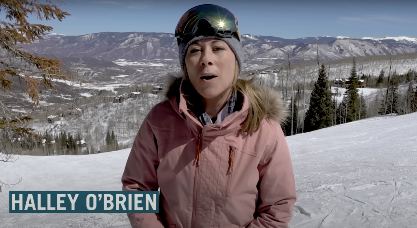 host speaking to camera with ski goggles on top of head and winter coat on, mountains sit behind her