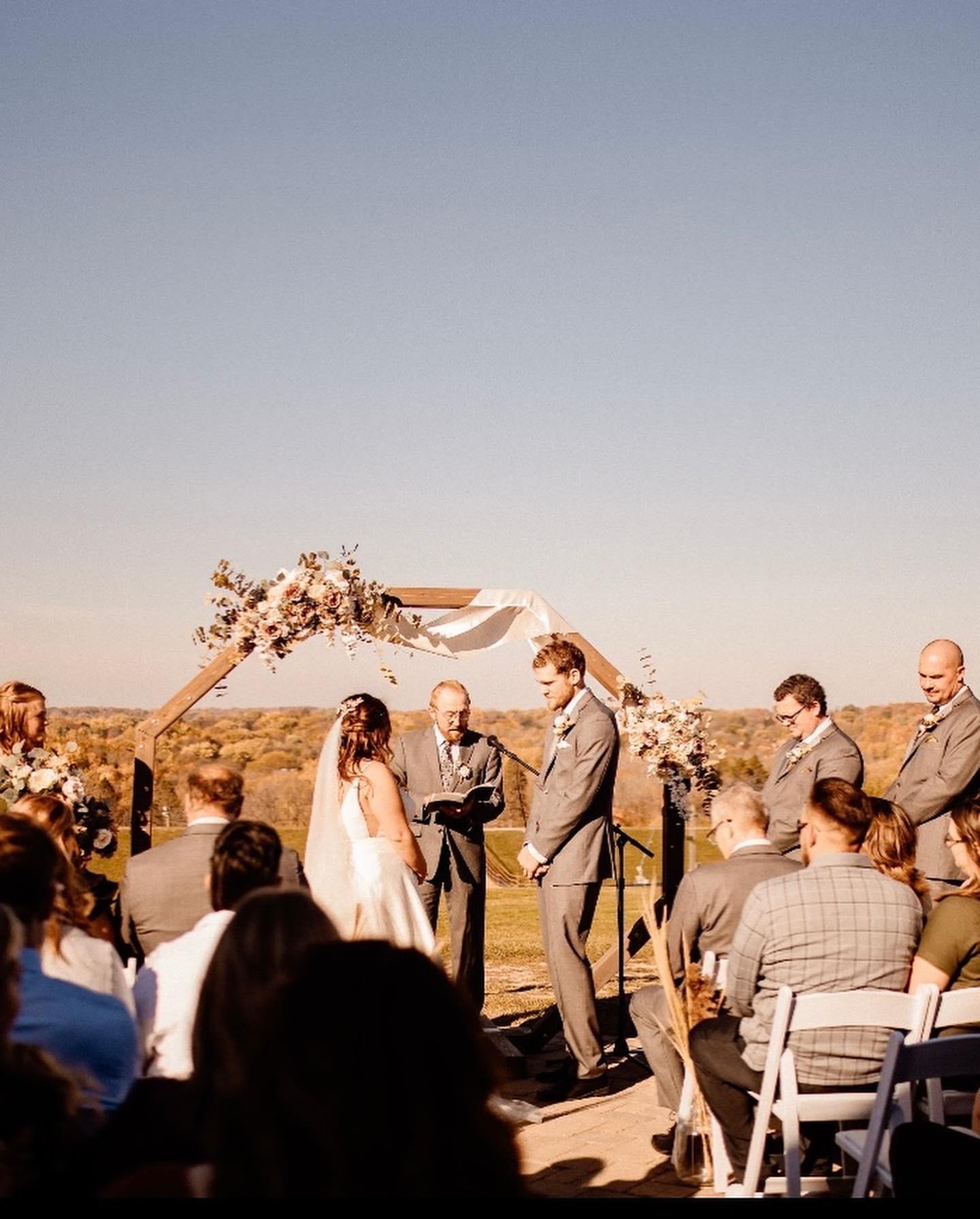 Outdoor wedding ceremony with couple under floral arch, guests seated.