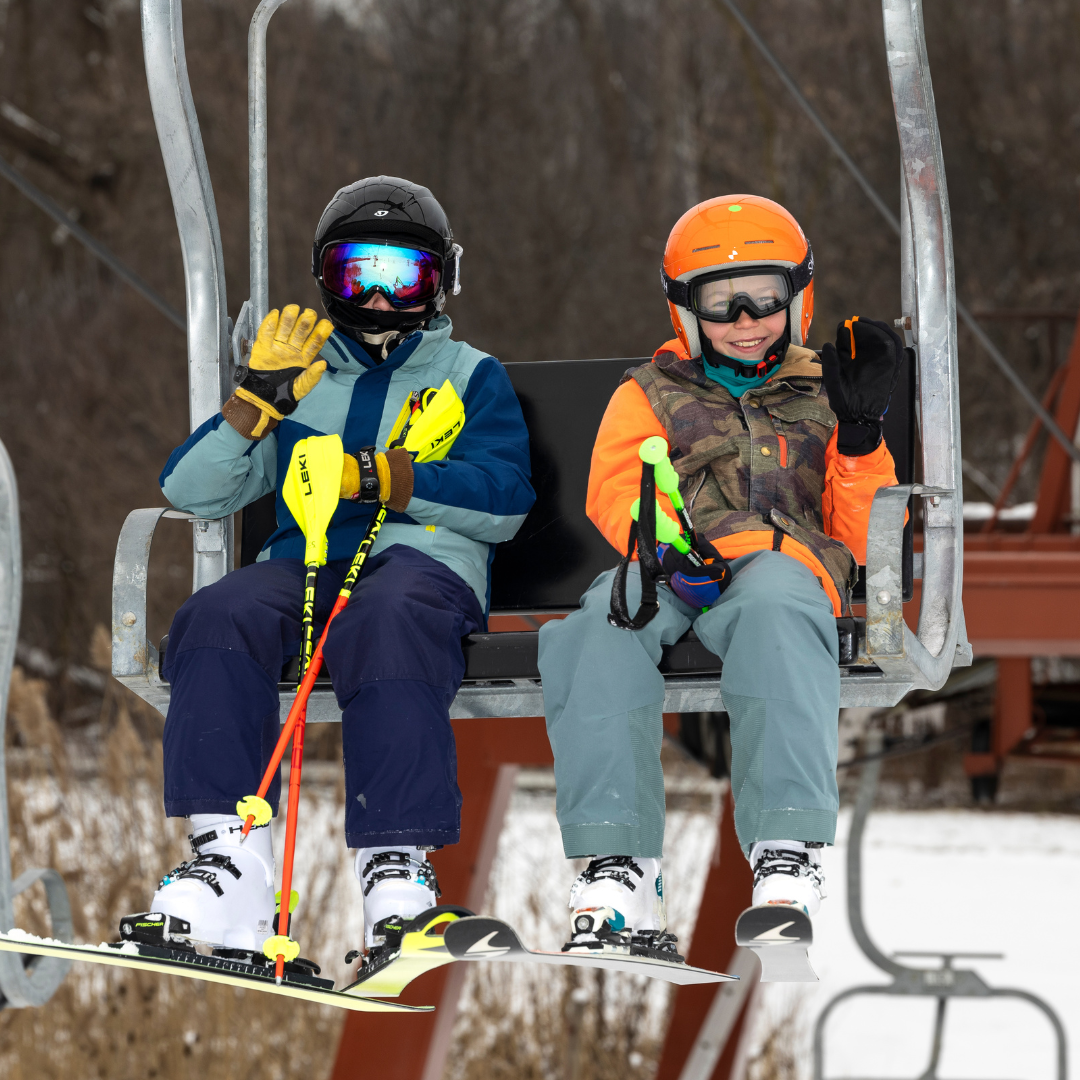 Two skiers in colorful gear wave from a ski lift.