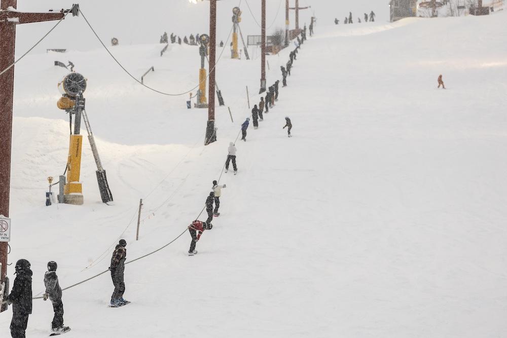 Skiers using a rope tow on a snowy slope, overcast sky.