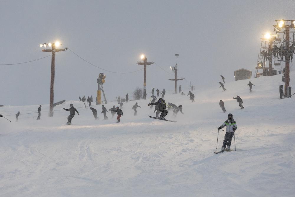 Skiers on a snowy slope under overcast sky with bright lampposts.