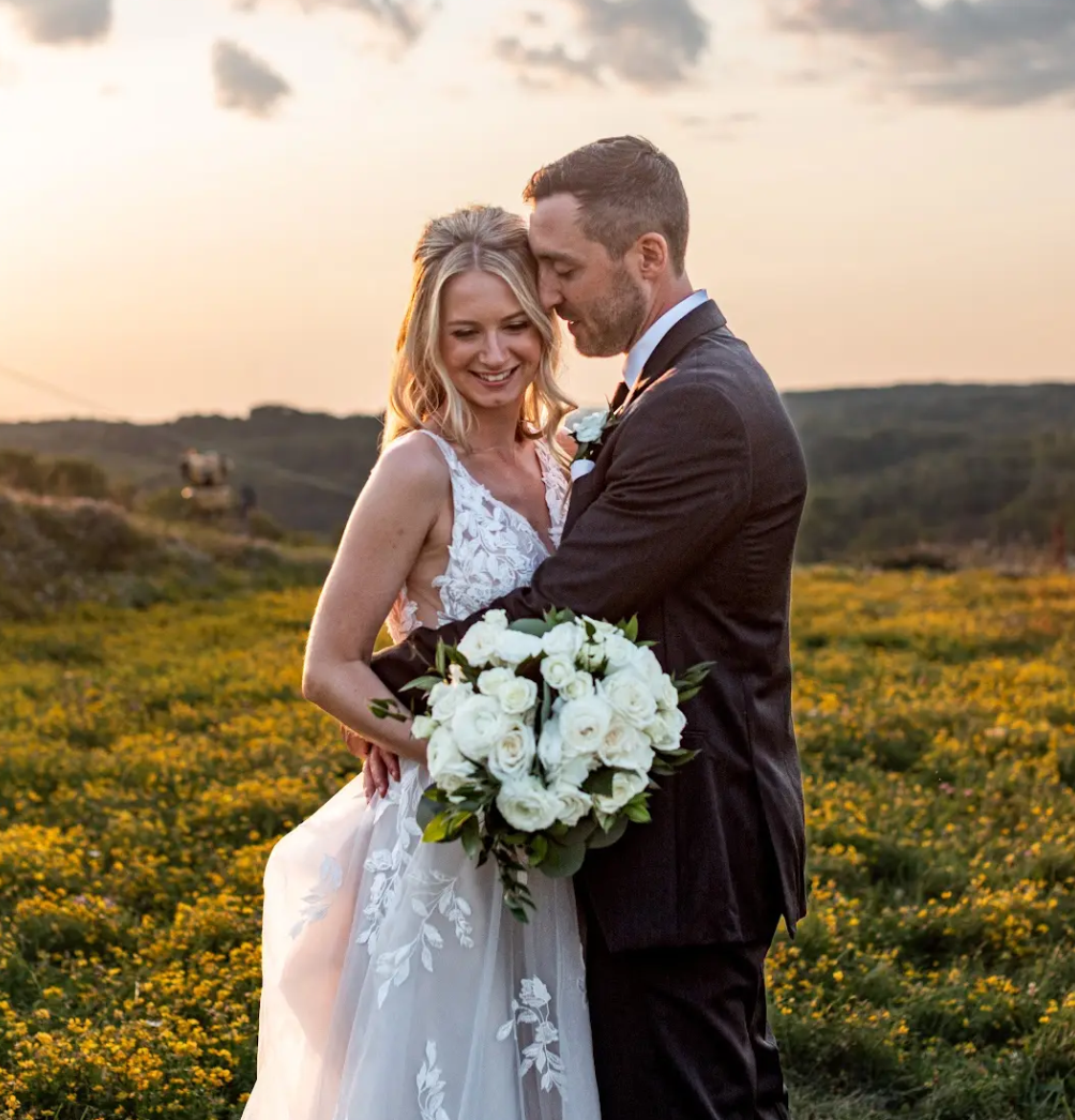 Bride and groom embracing in a field at sunset, holding a bouquet of white flowers.