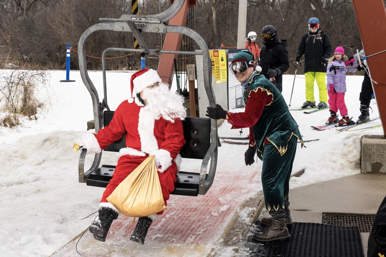 Santa Claus and a companion ride a ski lift as skiers wait nearby.