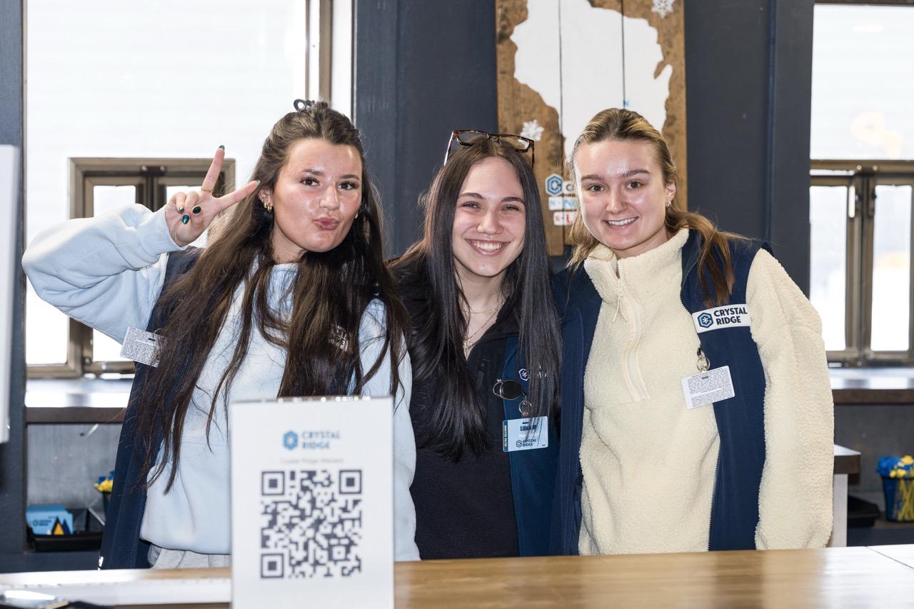 Three young women smiling behind a table with a QR code sign.