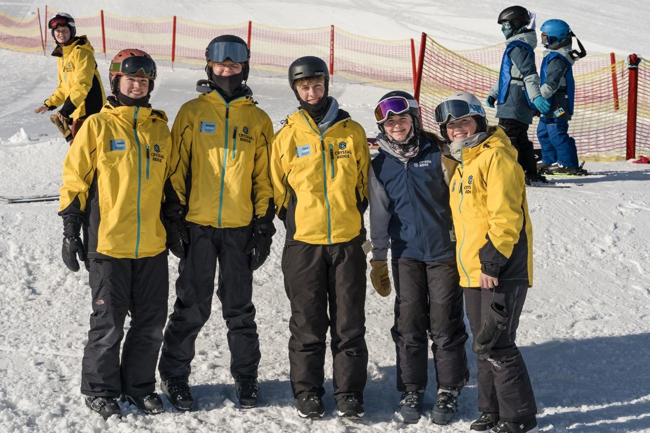 Six people in yellow jackets stand on a snowy mountain with climbers behind them.