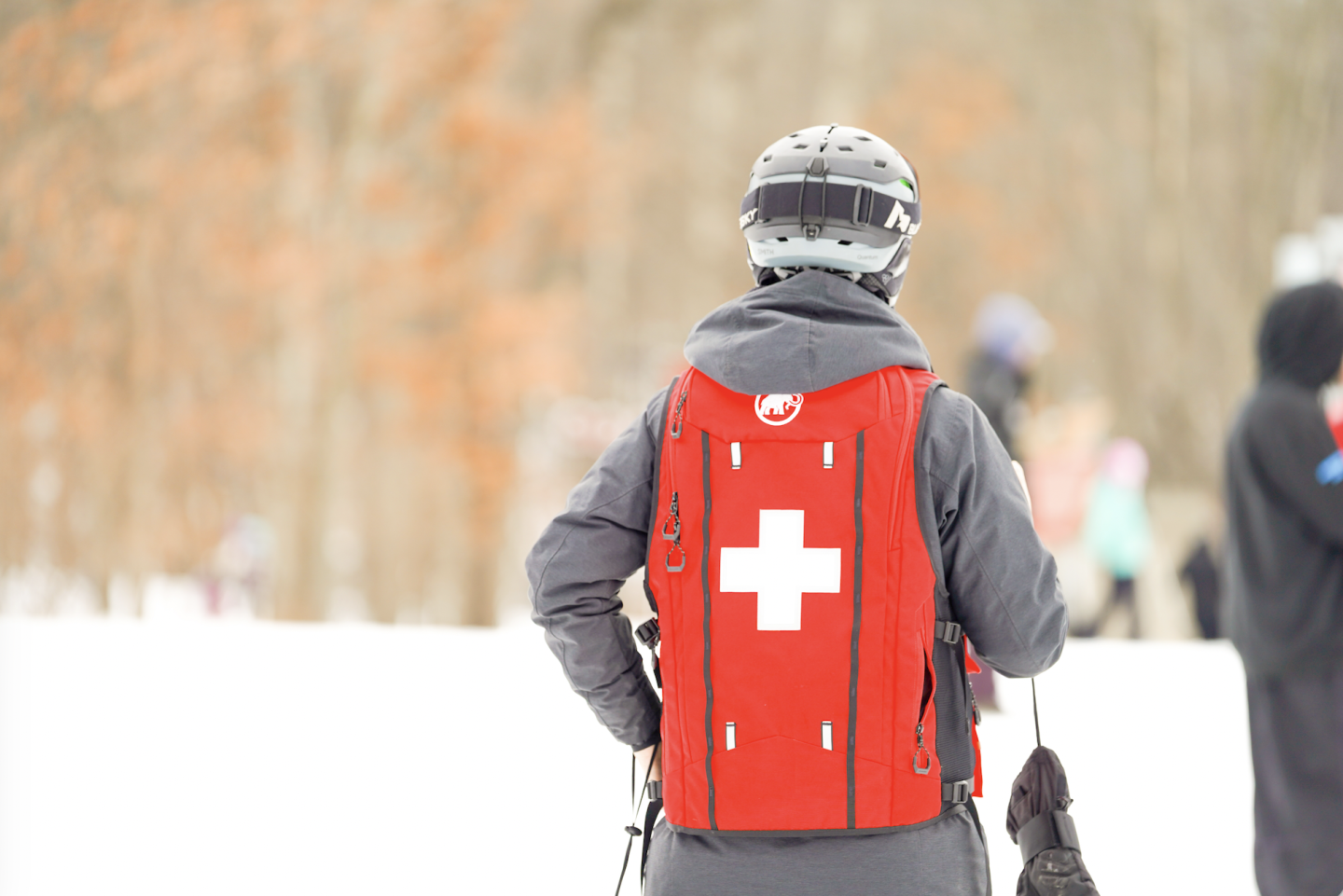 Ski patrol member in red vest with white cross standing on snowy slope.