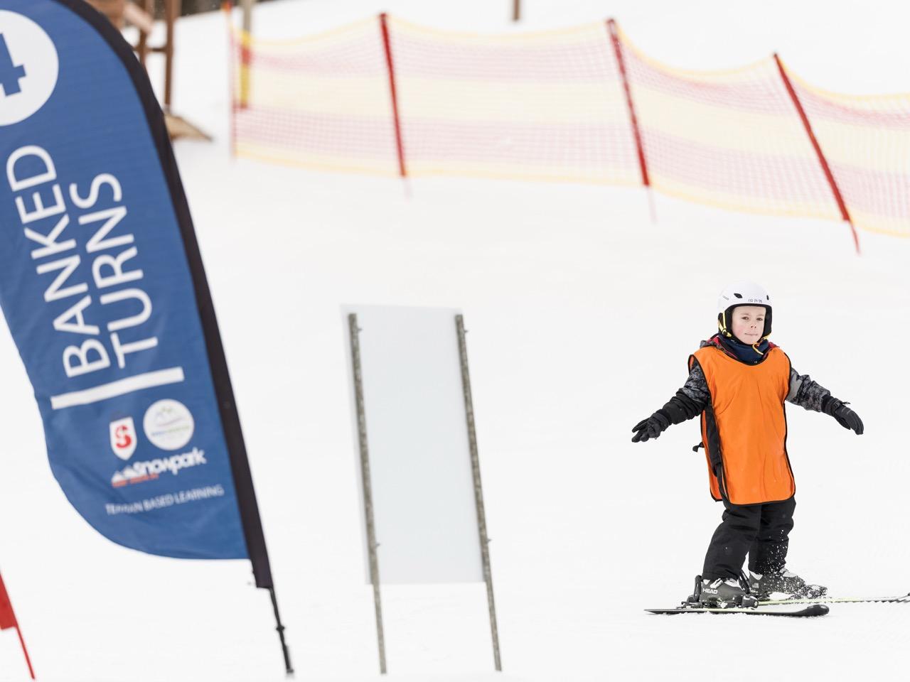 Snowboarder in orange jacket riding on a snowy slope near a “Banned Pitons” banner.
