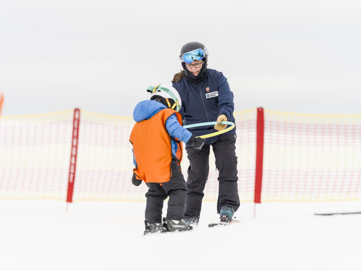 Adult helping a child ski on a snowy slope beside orange safety fencing.