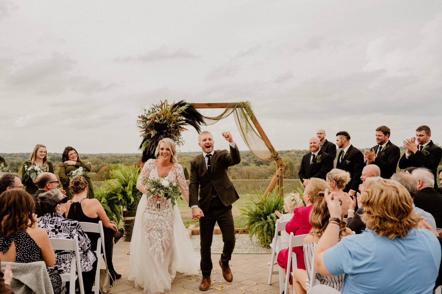 Bride and groom walk down an outdoor aisle as guests cheer.