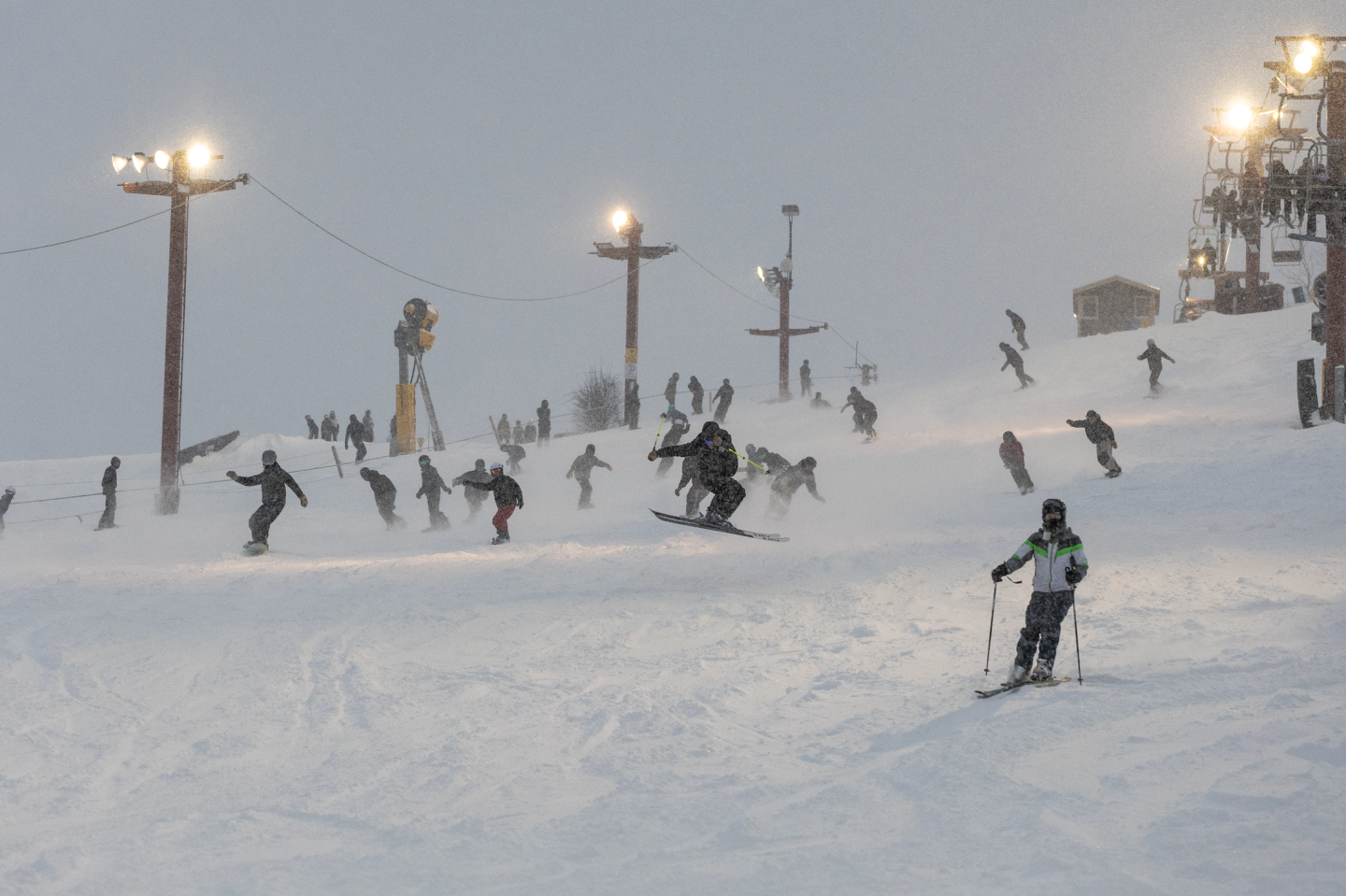 Skiers and snowboarders descend a snowy slope at dusk under bright lights.