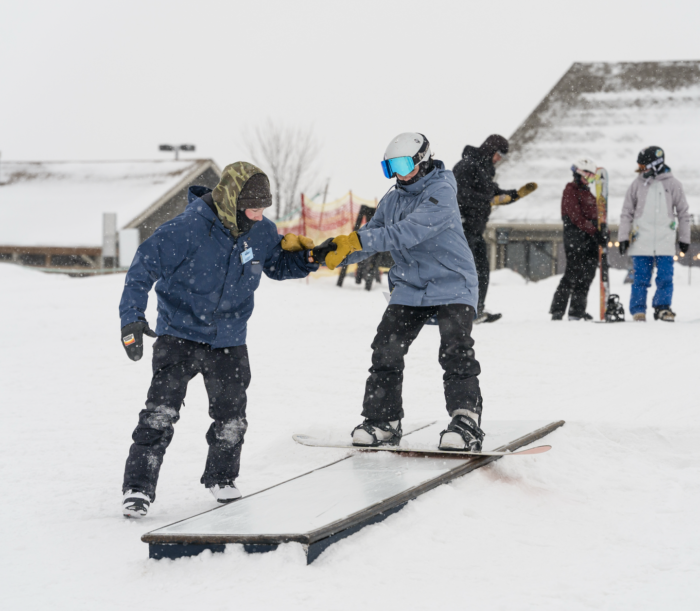 Snowboarders high-five near a rail in a snowy terrain park.