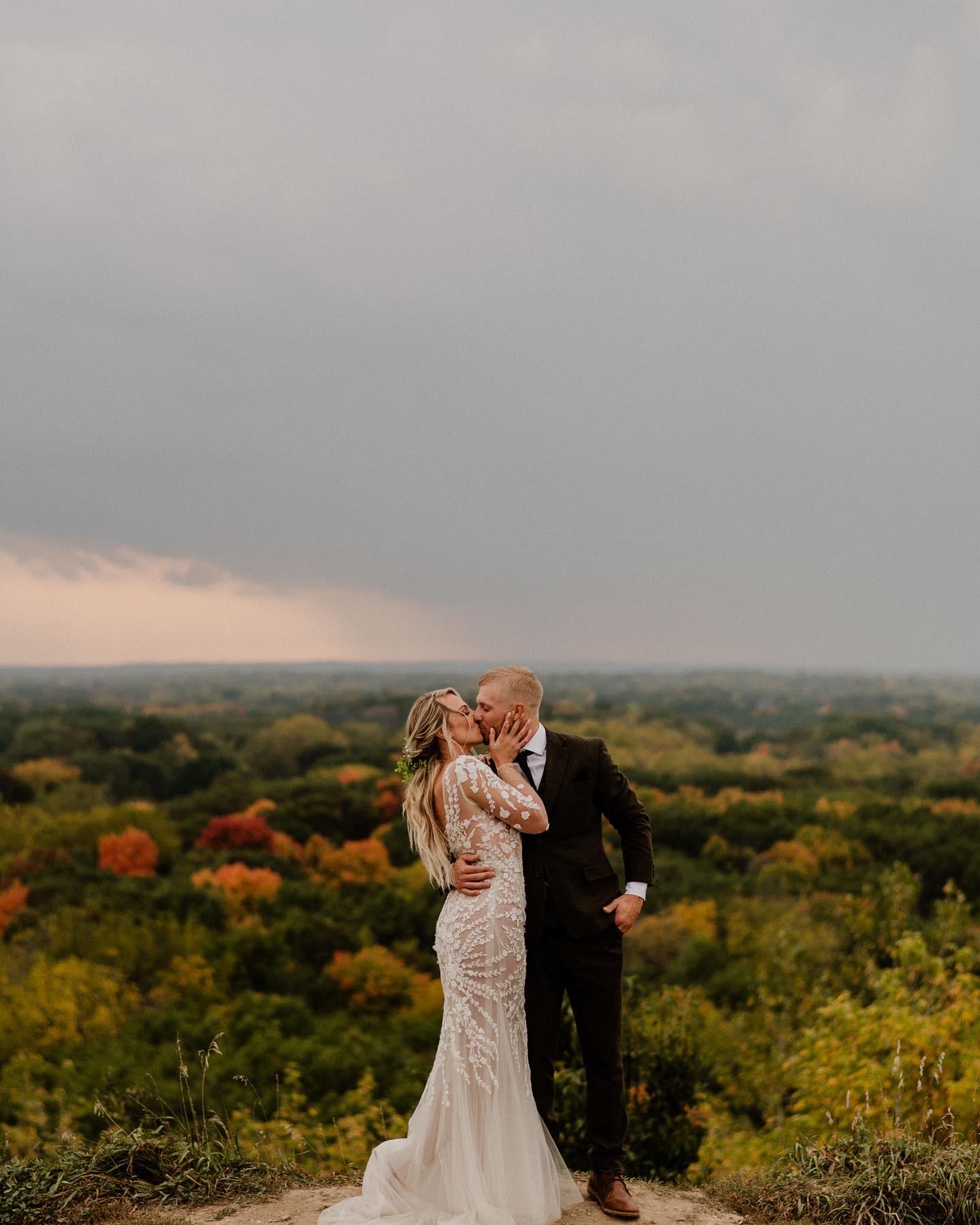 Bride and groom embracing on a hill overlooking autumn foliage under a cloudy sky.