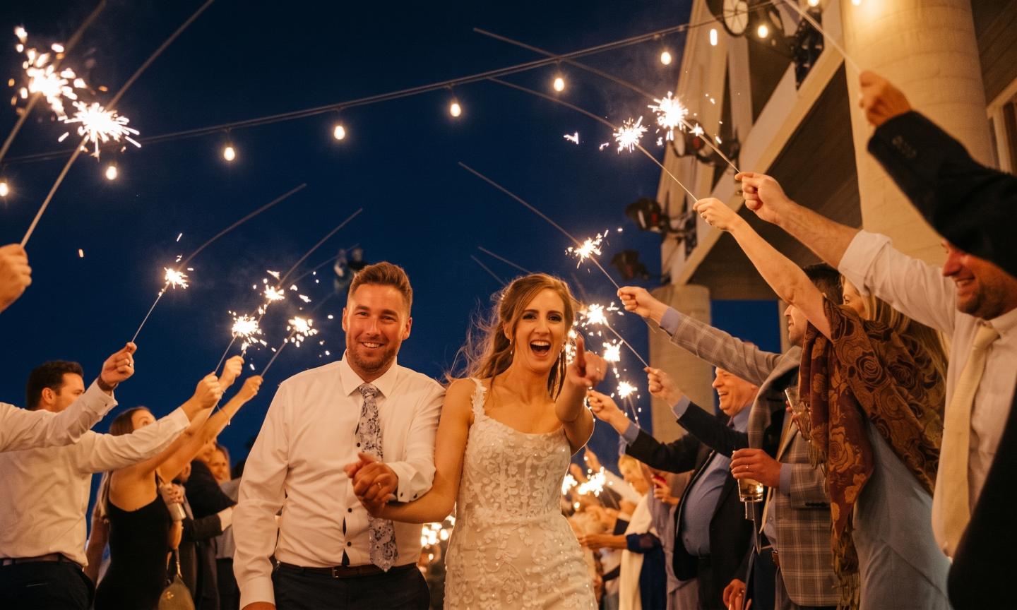 Bride and groom celebrate under string lights as guests wave sparklers.