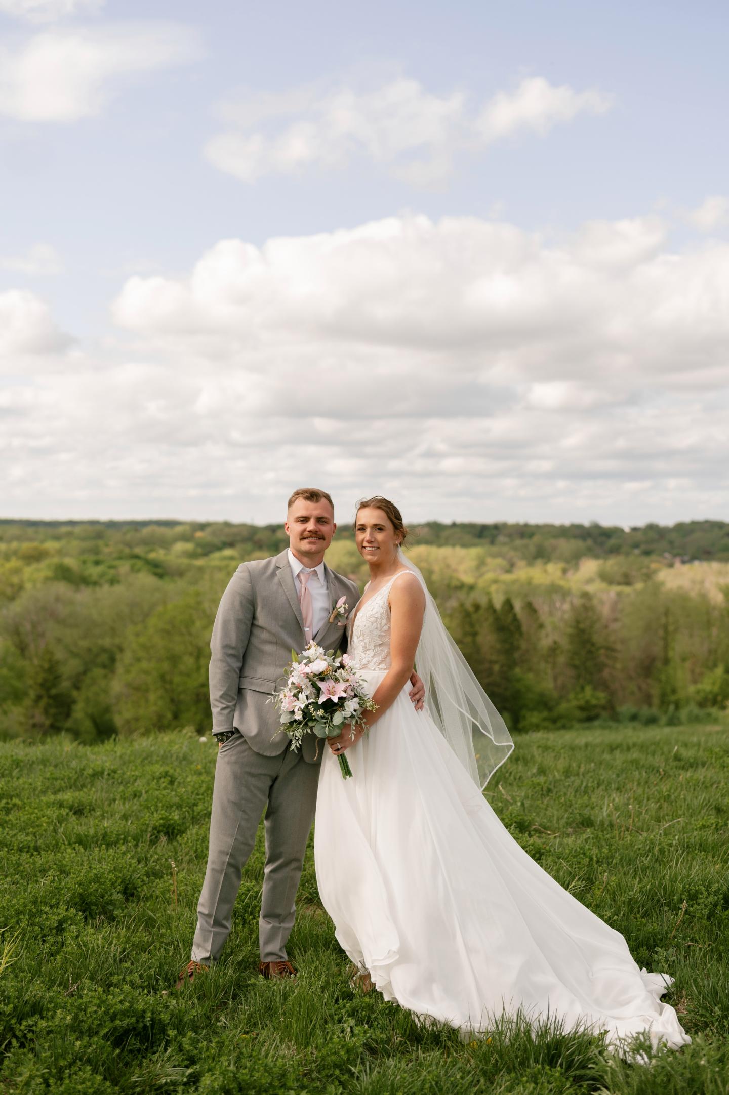 Bride and groom standing on a grassy hill with a forested landscape behind them.
