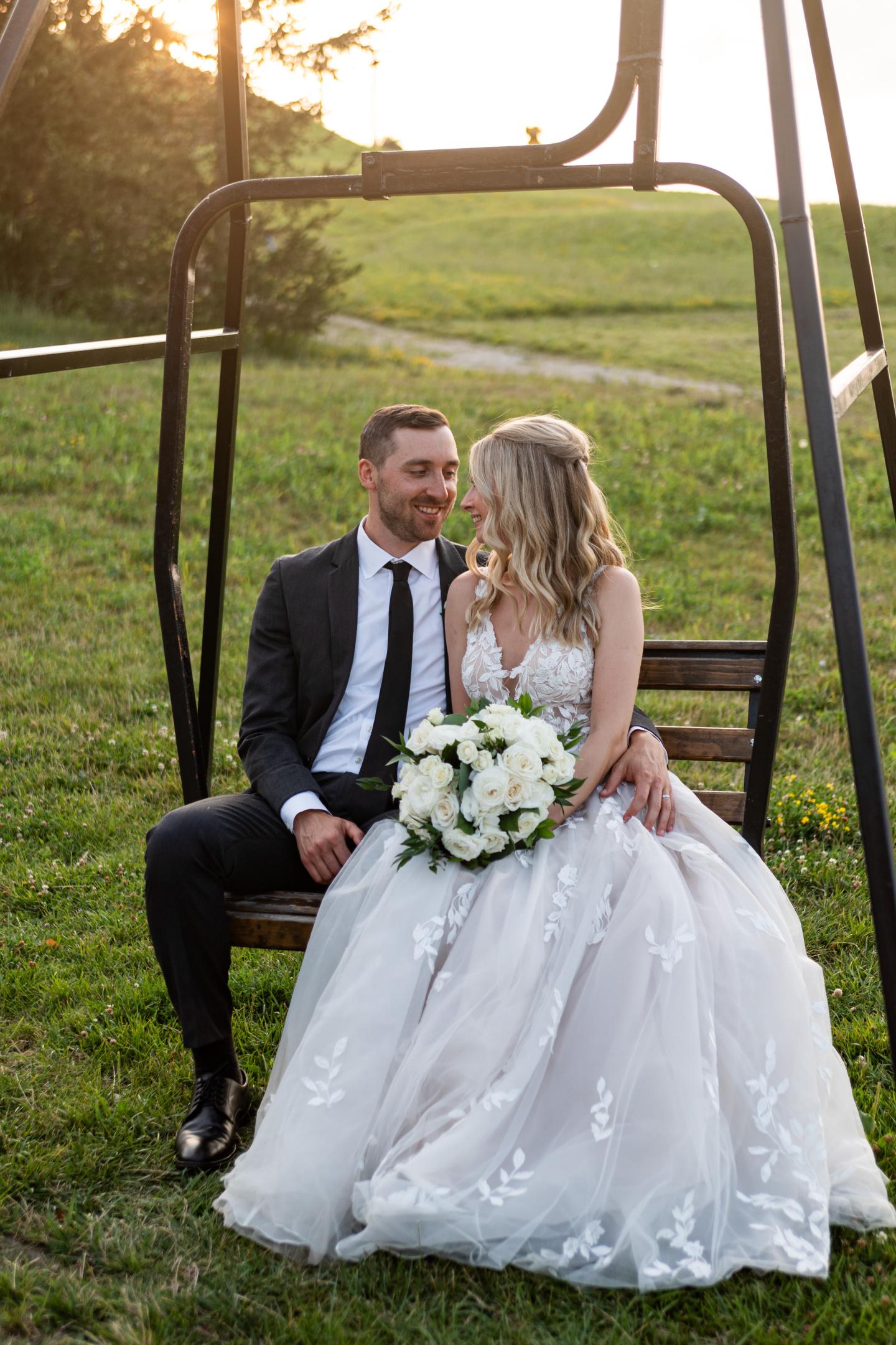 Bride and groom seated on a swing at sunset, holding a white bouquet.